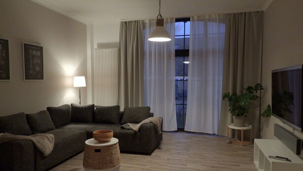 A modern living room with a sofa, a lamp, a potted plant, a TV, and a window with curtains. - Holiday Apartment "Am Conradhof" with Private Terrace and Wi-Fi (Goch - Pfalzdorf)