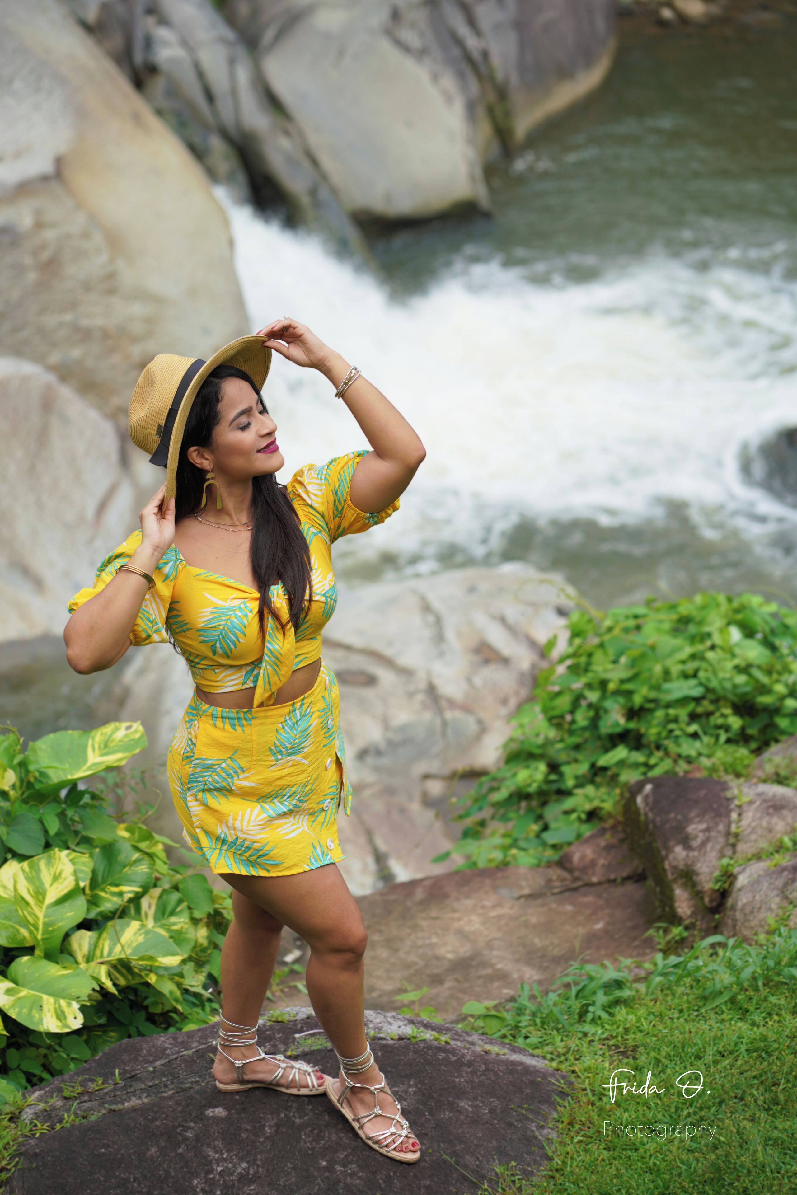 Una persona de pie sobre una roca junto a un río caudaloso, vestida con un atuendo amarillo y un sombrero.