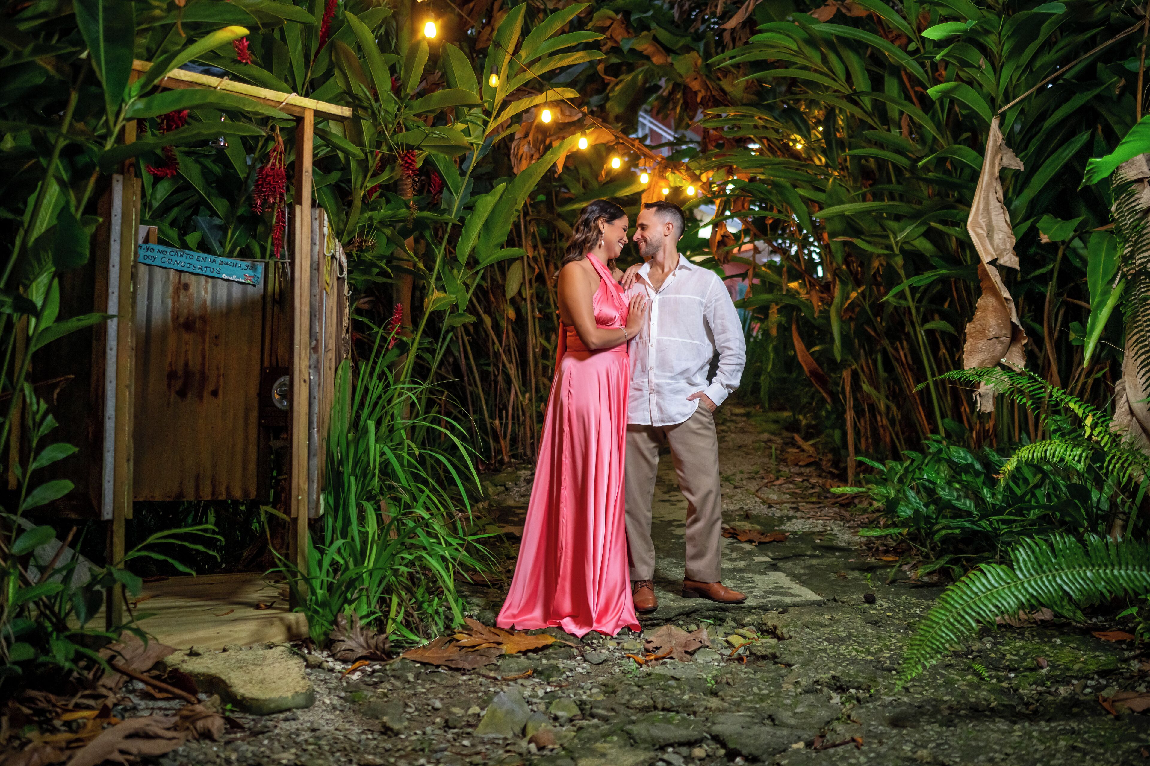 Un couple se tient dans un jardin luxuriant, avec une structure en bois et des guirlandes lumineuses.