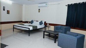 Une chambre d’hôtel comprenant un lit, un bureau, deux fauteuils, un ventilateur de plafond, un appareil de climatisation et un rideau épais.