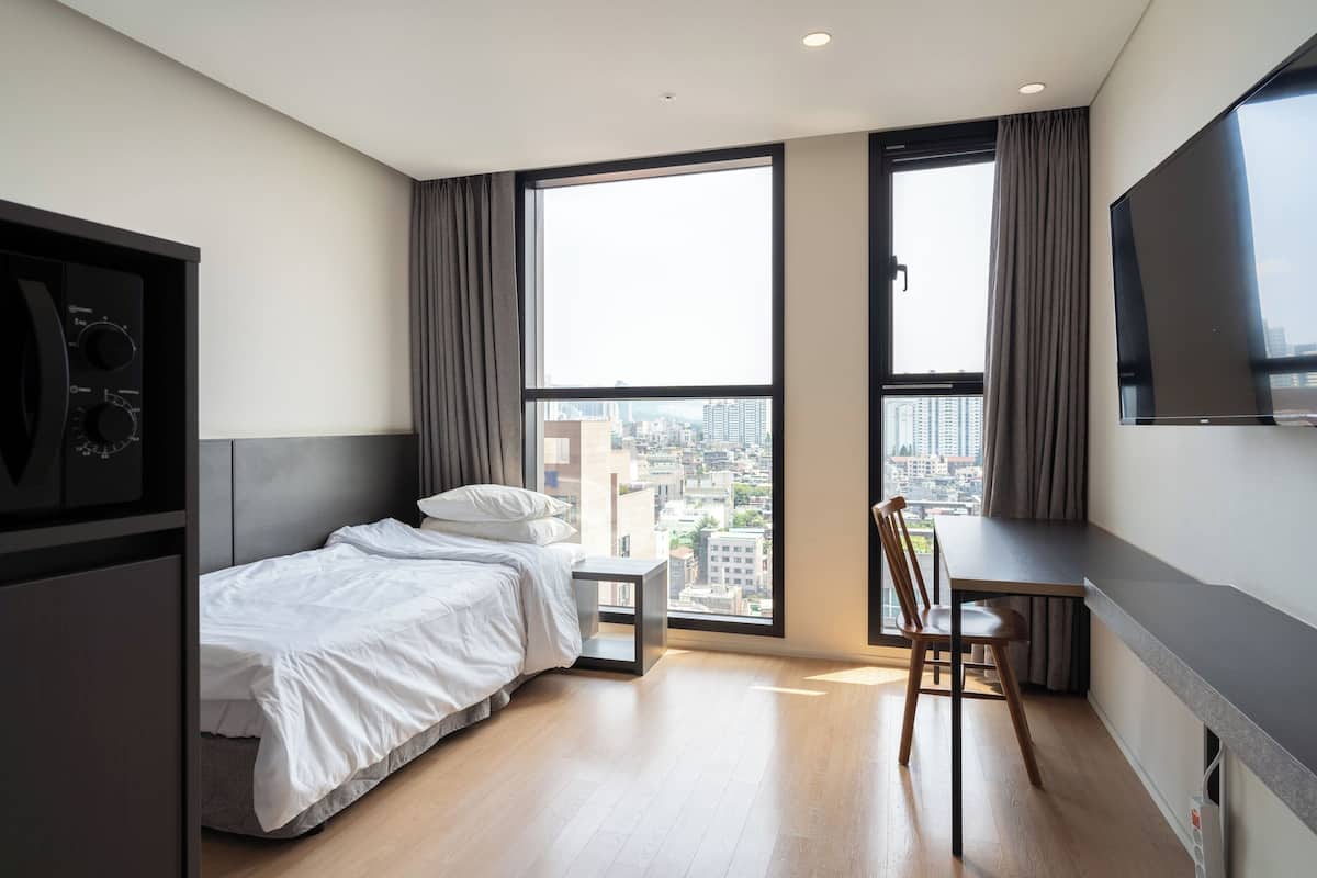 A modern hotel room with a bed, a small table, a chair, a TV, and large windows offering a city view.