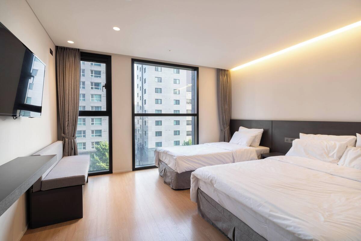 A hotel room with two beds, a desk, and a large window offering a city view.