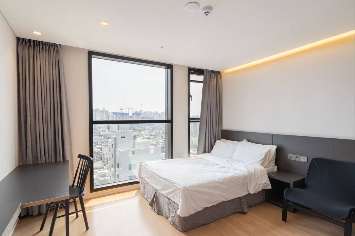 A modern hotel room with a large window offering a city view, a bed, a desk, and a chair.