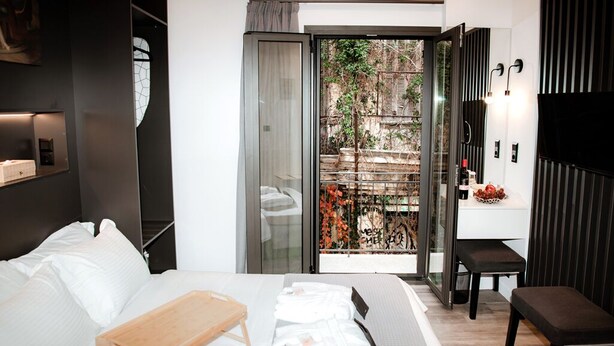 A modern hotel room with a bed, a wooden bedside table, a bench, a glass door leading to an outdoor area with a view of a garden and a building. - Athens Muses Suites (Athens)