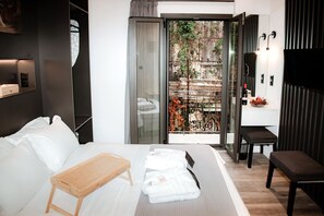 Ein modernes Hotelzimmer mit einem Bett, einem Nachttisch aus Holz, einer Bank und einer Glastür, die zu einem Außenbereich mit Blick auf einen Garten und ein Gebäude führt.