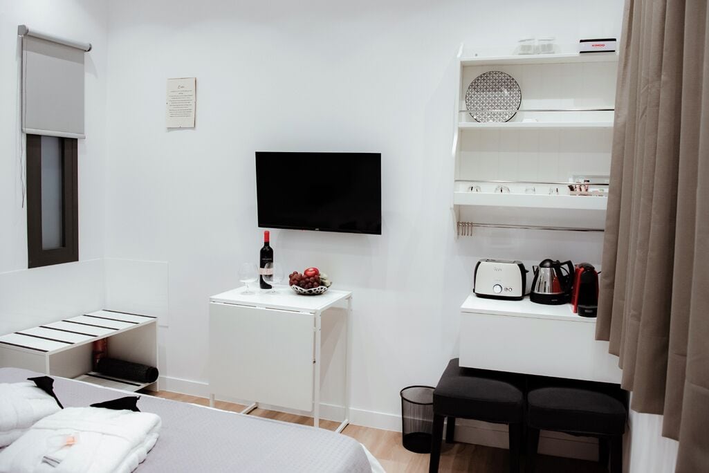 Una habitación de hotel moderna con cama, televisor, una pequeña mesa con una botella de vino y fruta, hervidor de agua y estante con artículos de tocador.