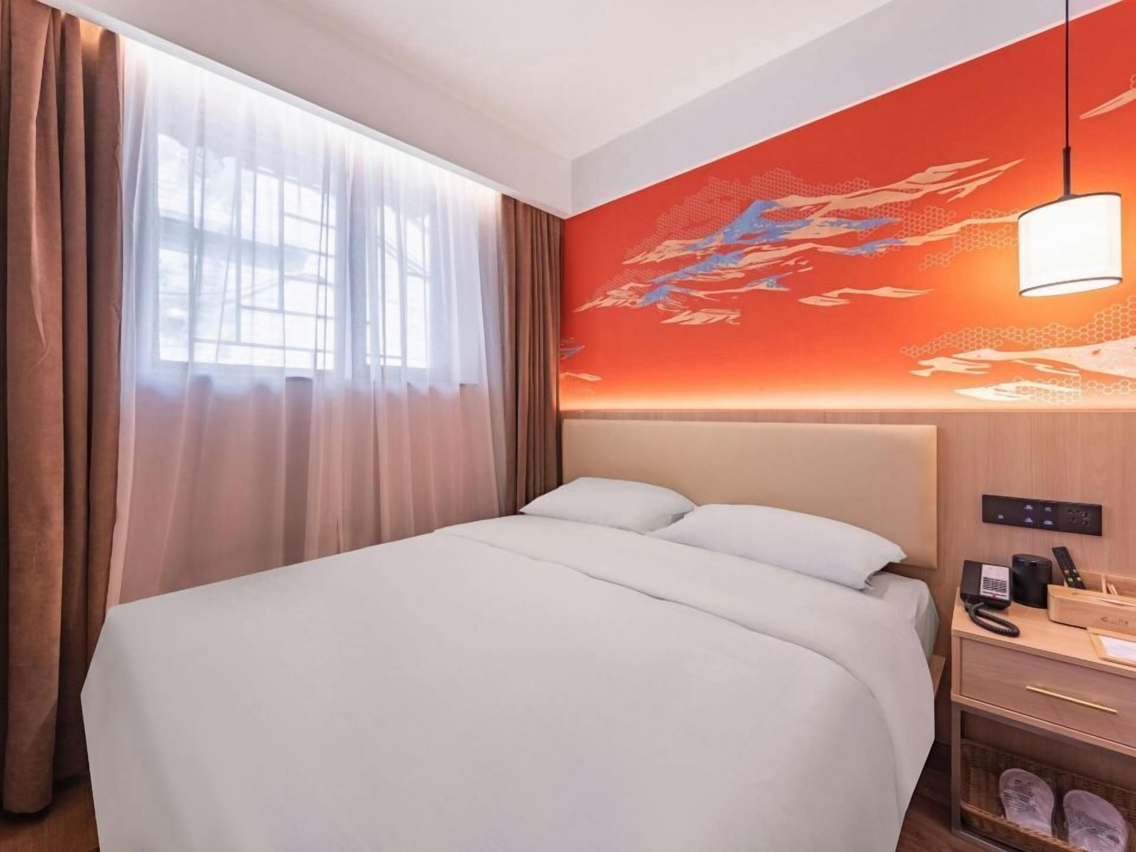 A hotel room with a large bed, a nightstand with a phone and remote controls, a wall-mounted lamp, and a window with sheer curtains.