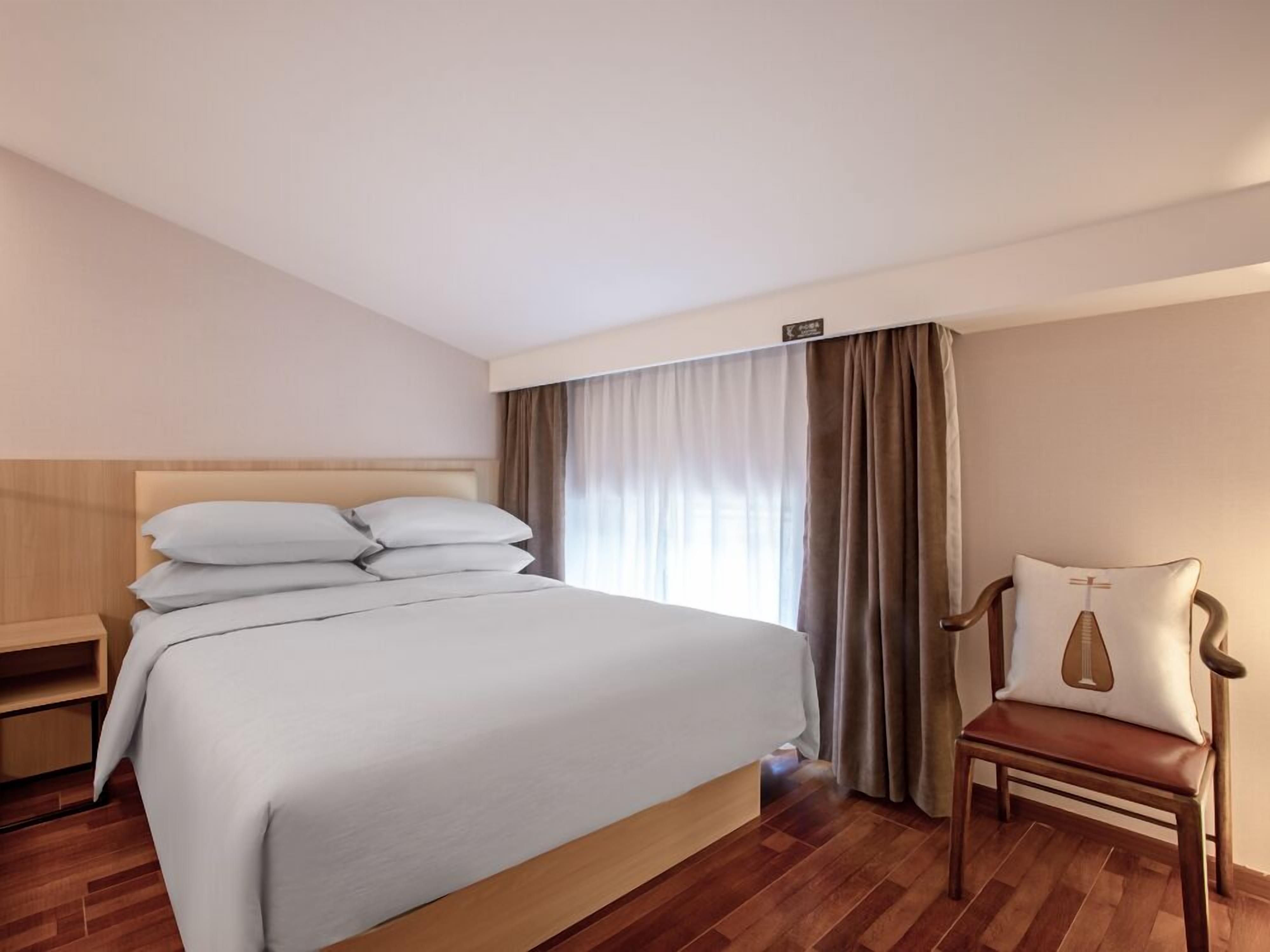 A hotel room with a large bed, a wooden nightstand, a chair, and a window with curtains.