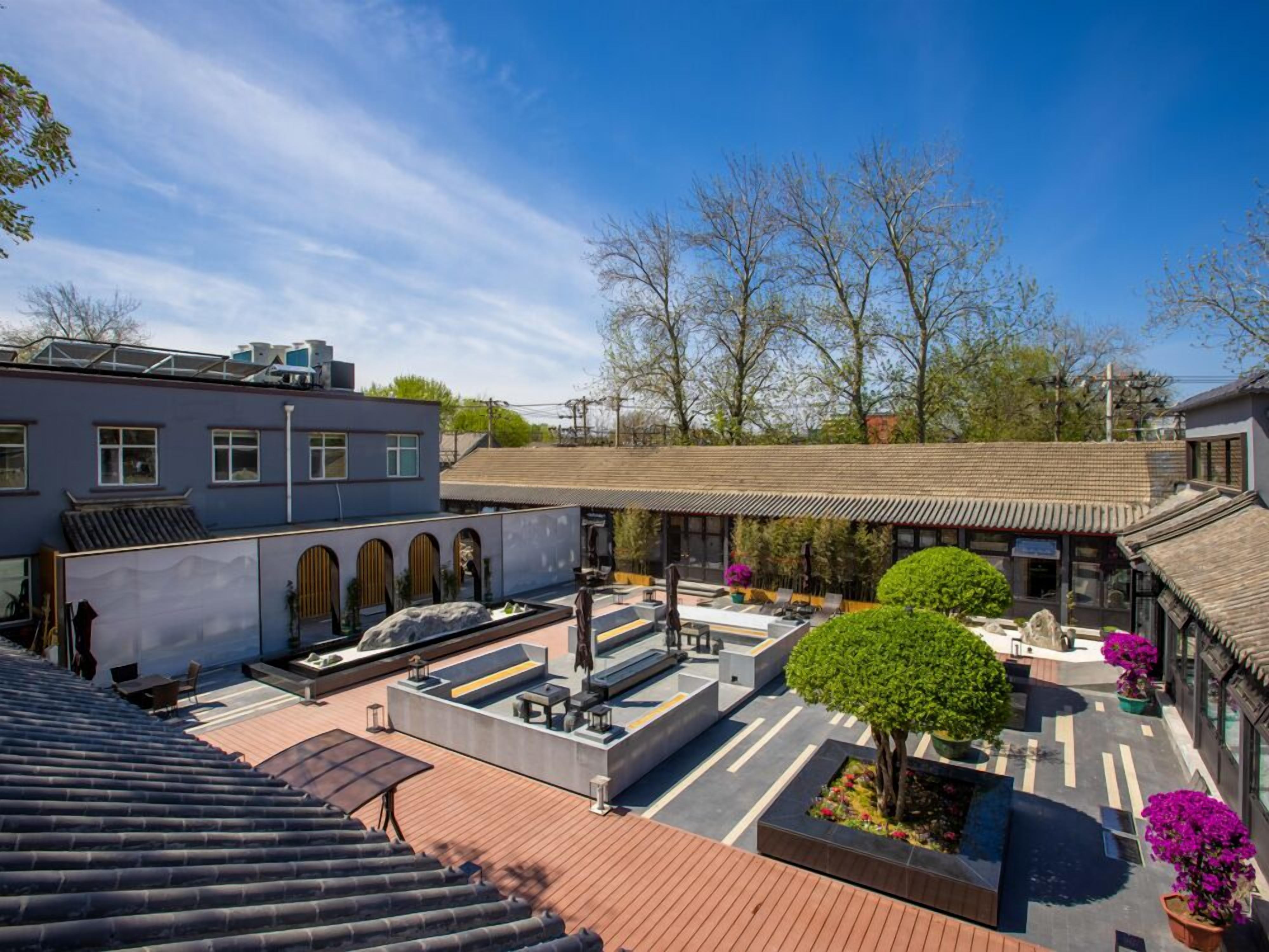 Une cour comprenant un bâtiment de style traditionnel, une structure moderne et un petit jardin agrémenté d’un arbre et de plantes en pot.