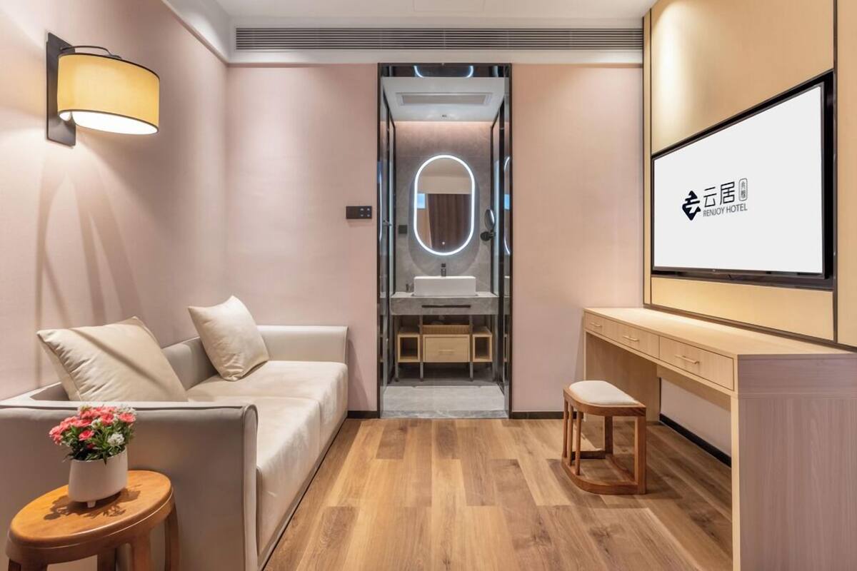 Ein modernes Hotelzimmer mit einer Couch, einem kleinen Tisch mit Blumen, einem Schminktisch mit Spiegel und einem Fernseher, der den Namen des Hotels zeigt.