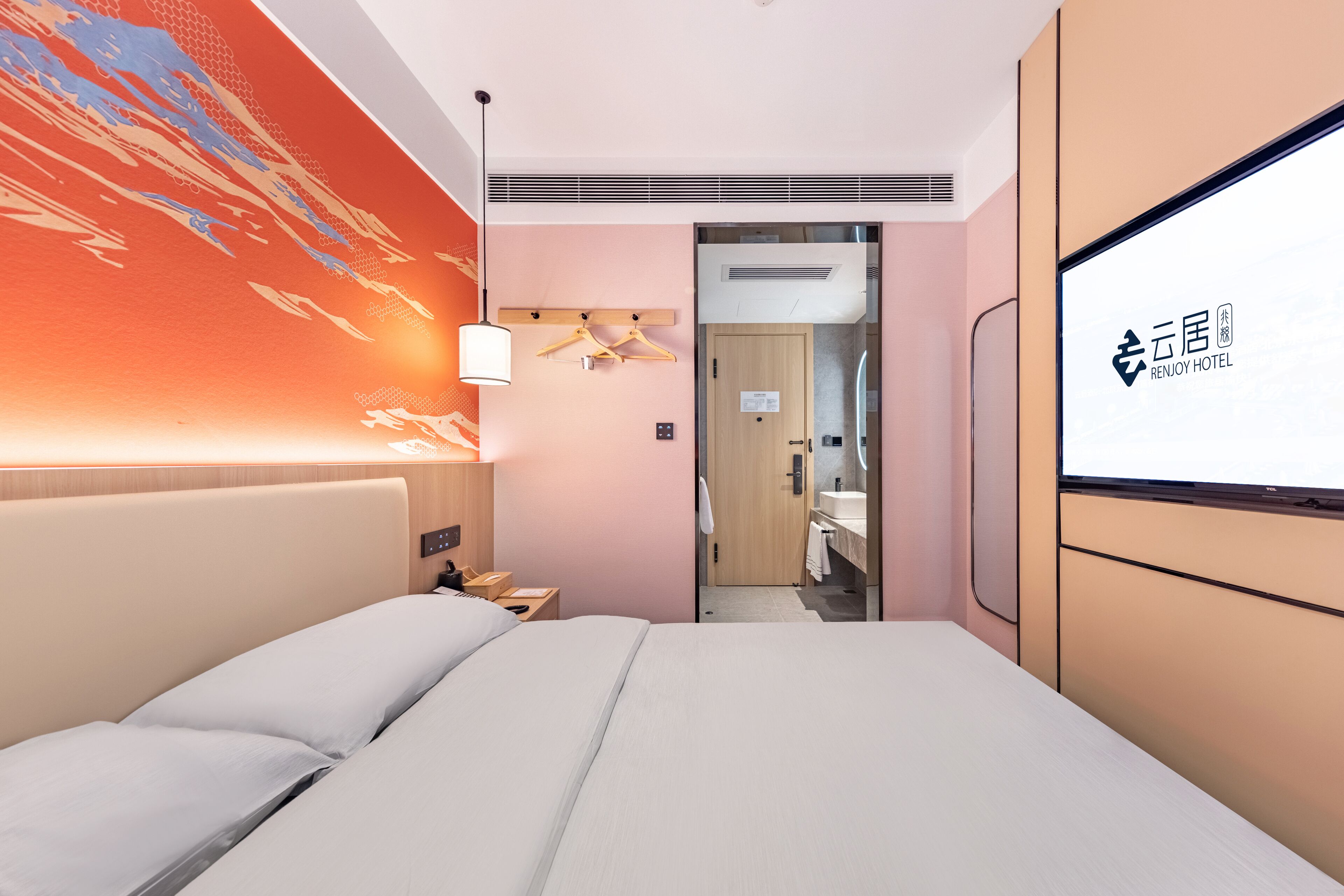 Une chambre d’hôtel comprenant un lit, un téléviseur fixé au mur, une porte donnant accès à une salle de bain et un mur orné d’un motif géométrique.