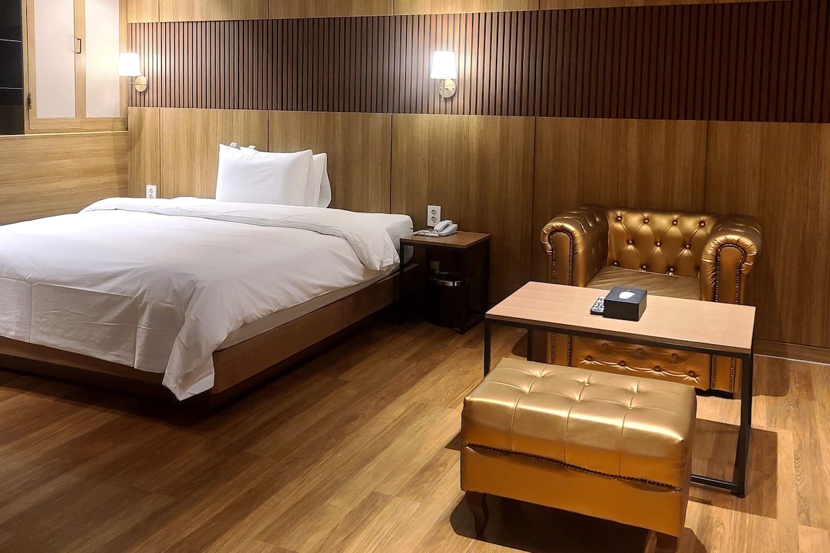 A modern hotel room with a wooden panel wall, a large bed, a nightstand, a leather armchair, and a small table.