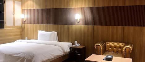A modern hotel room with a wooden panel wall, a large bed, a nightstand, a leather armchair, and a small table.