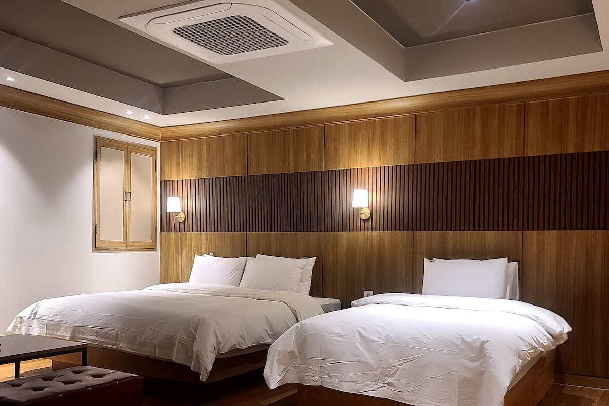 A modern hotel room with two beds, wooden paneling, a bench, and a ceiling vent.