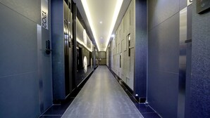A modern hallway with a long, straight layout, featuring a digital elevator display, a door with a number sign, and a clean, tiled floor. - G&G Hotel (Ulsan)