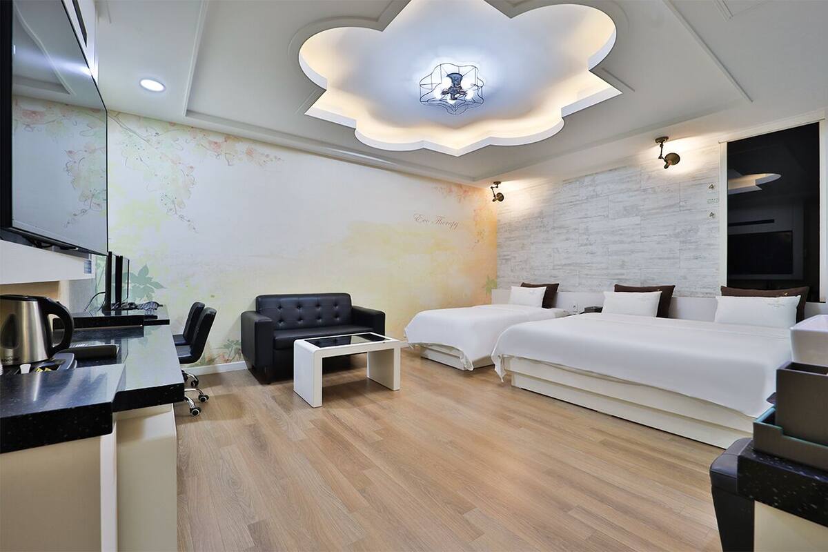 A modern hotel room with a wooden floor, a black sofa, a white bed, and a desk with a kettle.