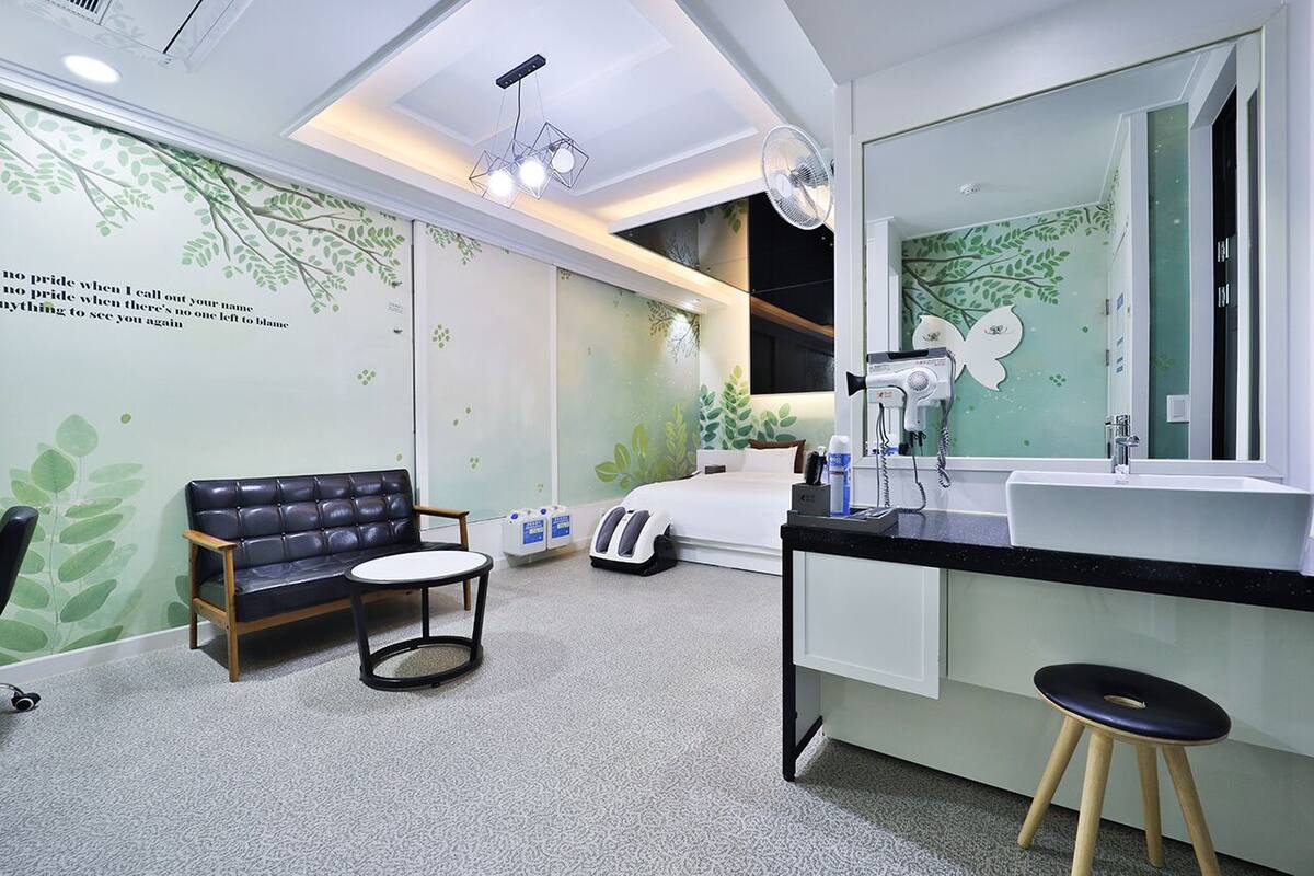 A modern hotel room with a bed, a sofa, a small table, a desk with a mirror, and a bathroom with a sink and mirror.