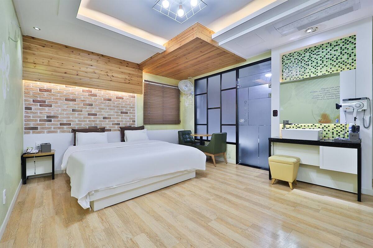 A modern hotel room with a large bed, a wooden ceiling, a brick accent wall, a bathroom with a mirror and sink, and a small table with a chair.