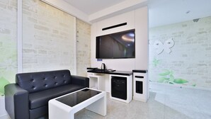 A modern living room with a black leather sofa, a glass coffee table, a flat-screen TV mounted on the wall, and a kitchen area with a black countertop and white cabinets. - G&G Hotel (Ulsan)