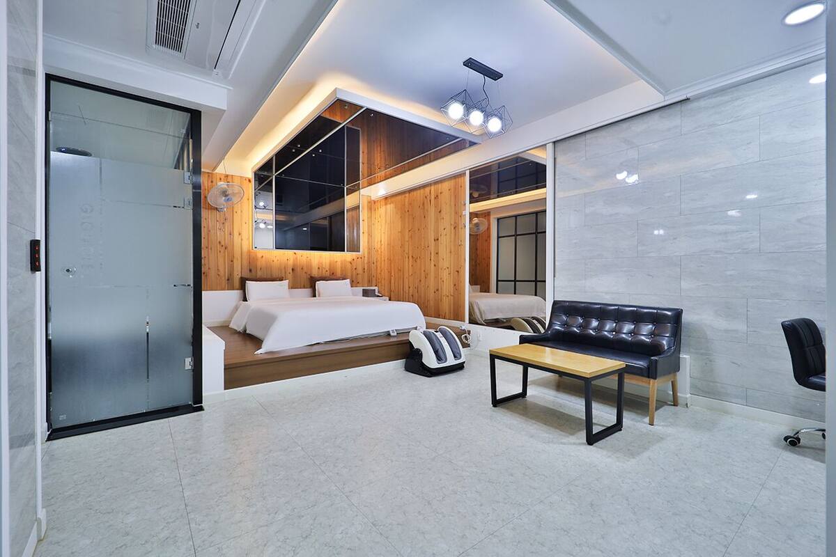 A modern hotel room with a large bed, a massage chair, a wooden accent wall, and a large window.