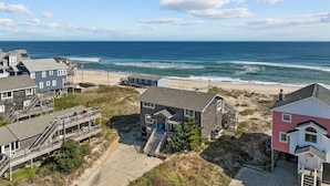 A coastal property with multiple buildings, including a house with a porch and a staircase leading to a deck, situated on a sandy beach with the ocean in the background. - 7019- Sara's Sea Breeze - Oceanfront Rodanthe | Hot Tub | Sleeps 12 (Rodanthe)