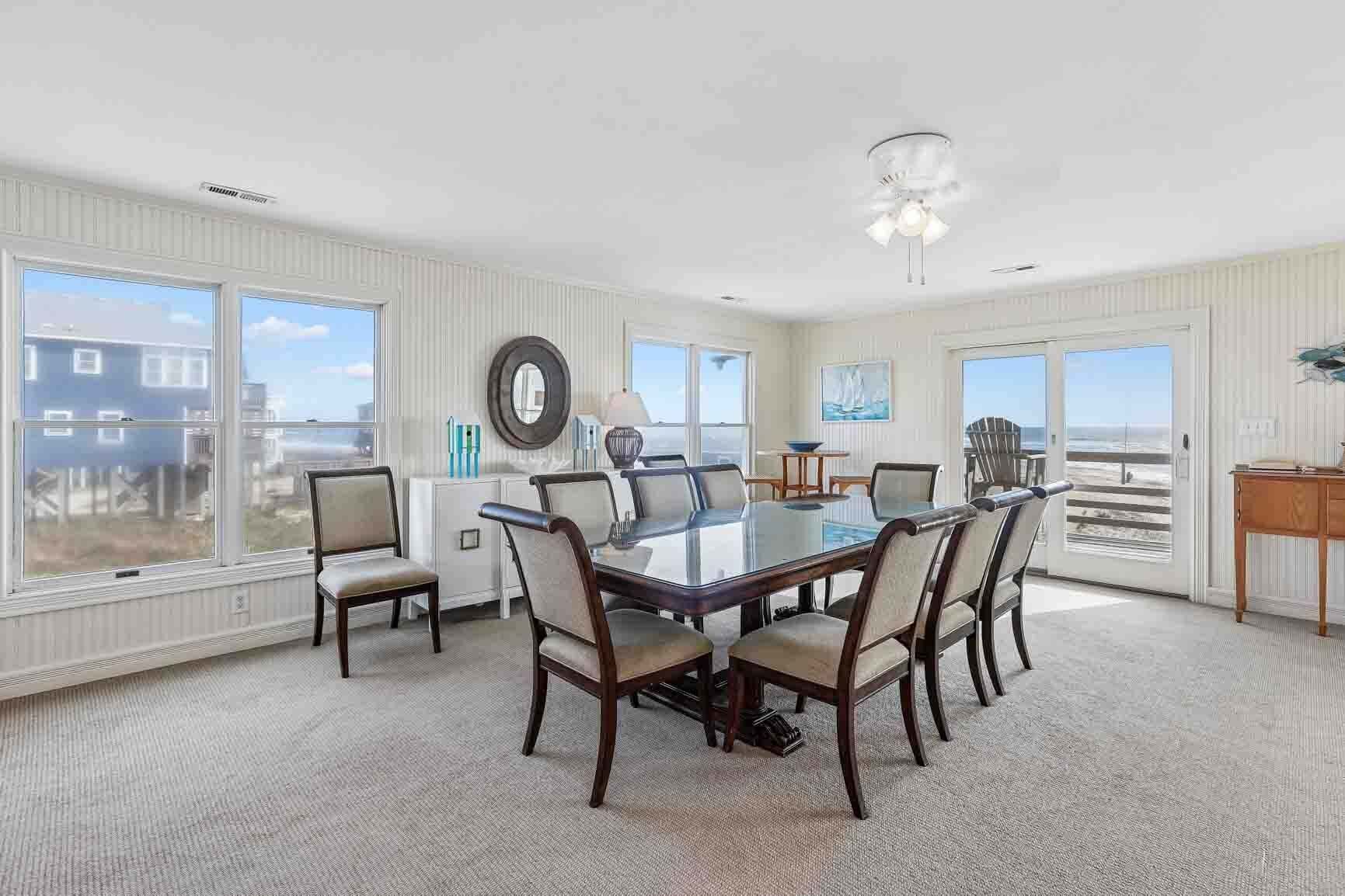 A spacious dining room with a large table and chairs, large windows, and a view of the beach.