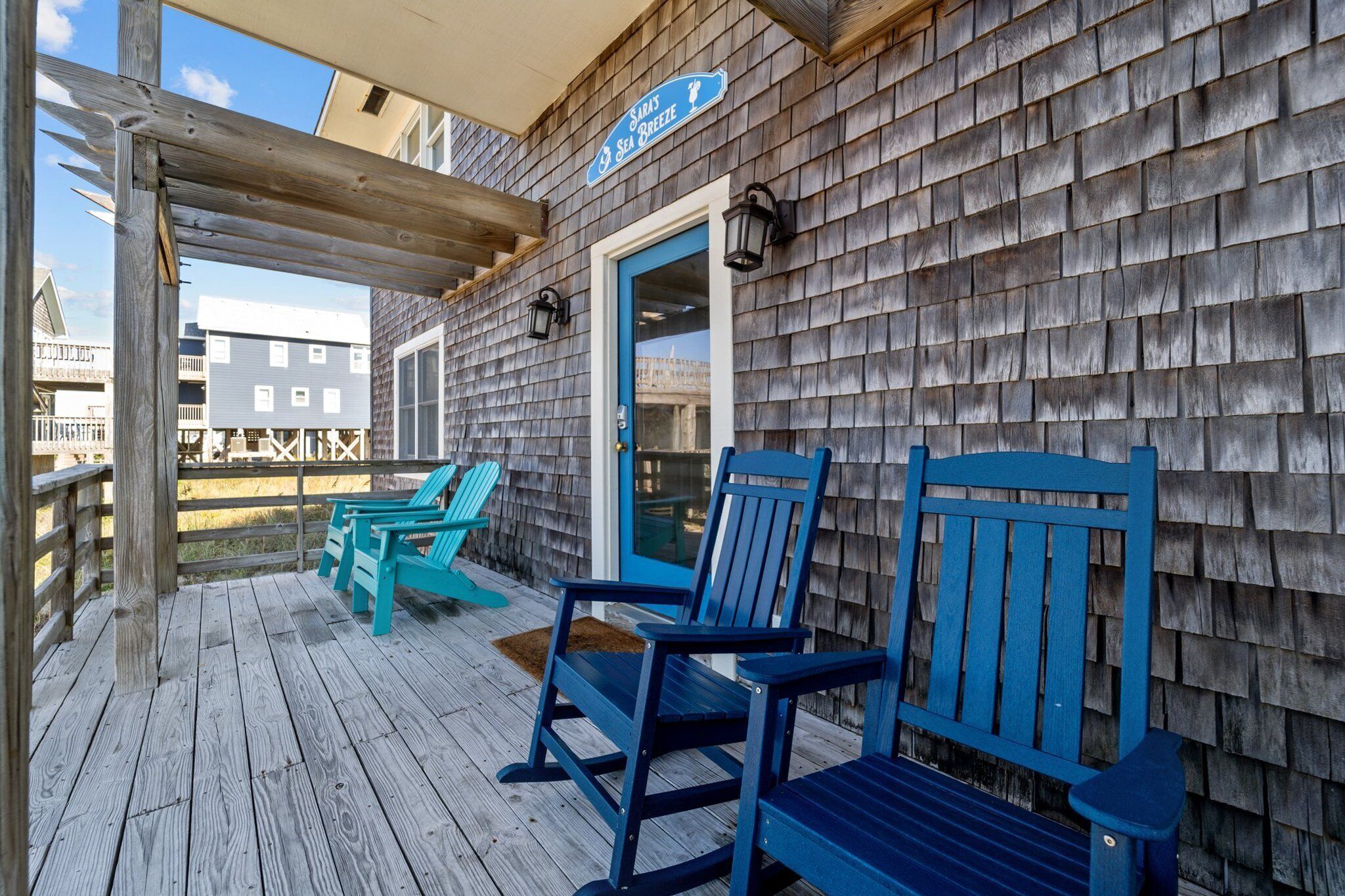 A wooden deck with blue Adirondack chairs and a blue bench, a wooden pergola, and a sign reading "Saras Sea Breeze".