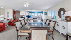 A spacious dining area with a large table and chairs, a kitchen in the background, and a view of the outdoors. - 7019- Sara's Sea Breeze - Oceanfront Rodanthe | Hot Tub | Sleeps 12 (Rodanthe)