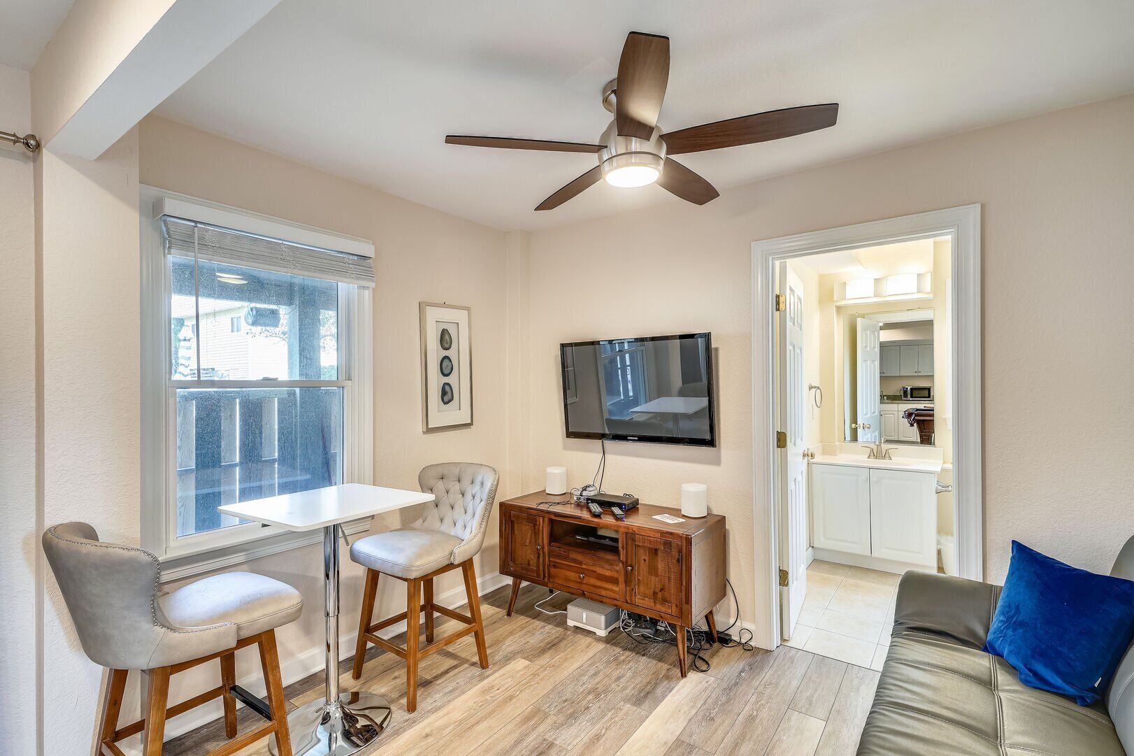 Un salon moderne doté d’un ventilateur de plafond, d’un téléviseur à écran plat fixé au mur, d’un coin repas avec une table et des tabourets de bar, et offrant une vue sur la cuisine grâce à une porte ouverte.