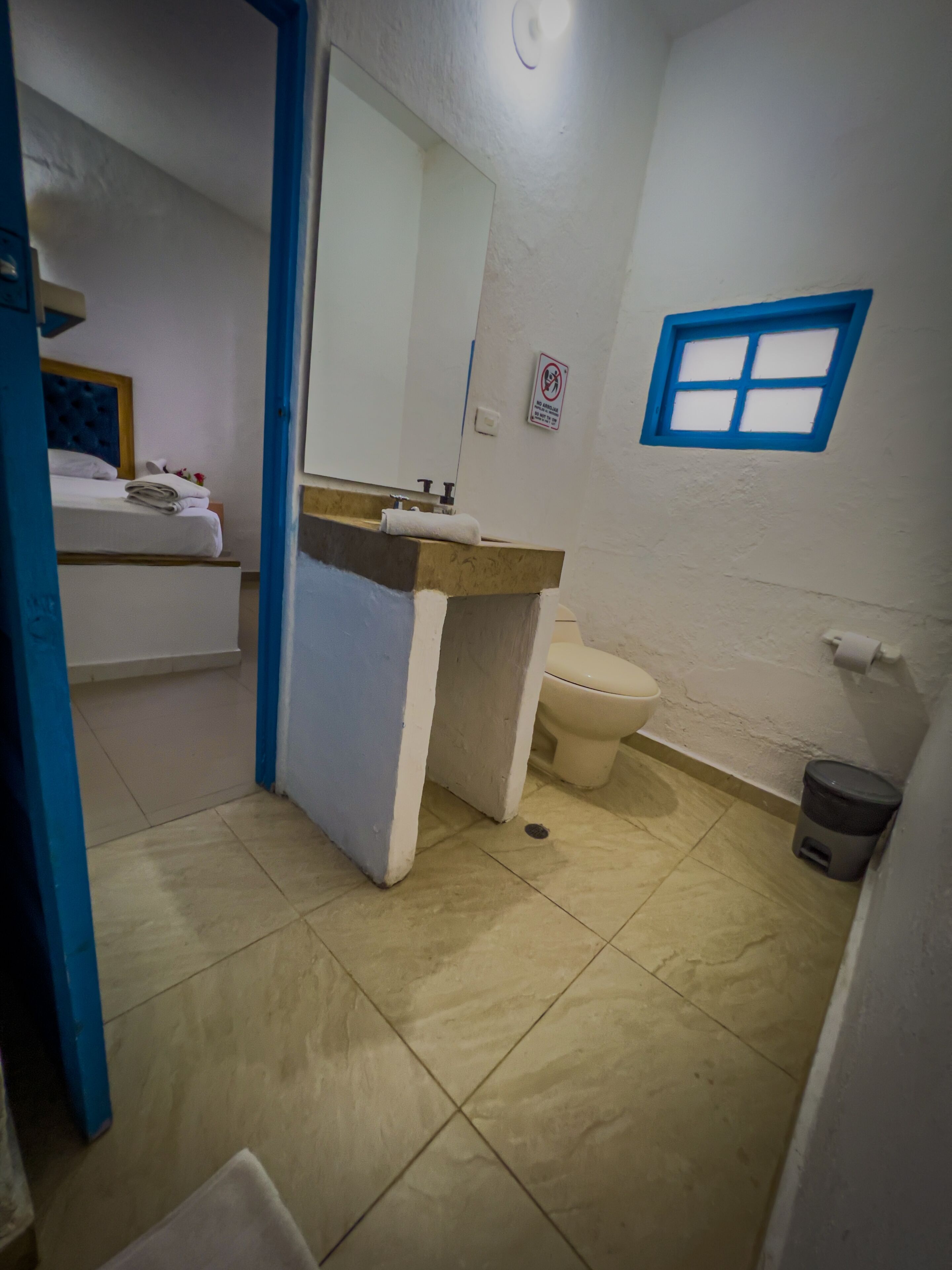 A small bathroom with a toilet, sink, and a mirror. There is a blue-framed window and a sign on the wall.