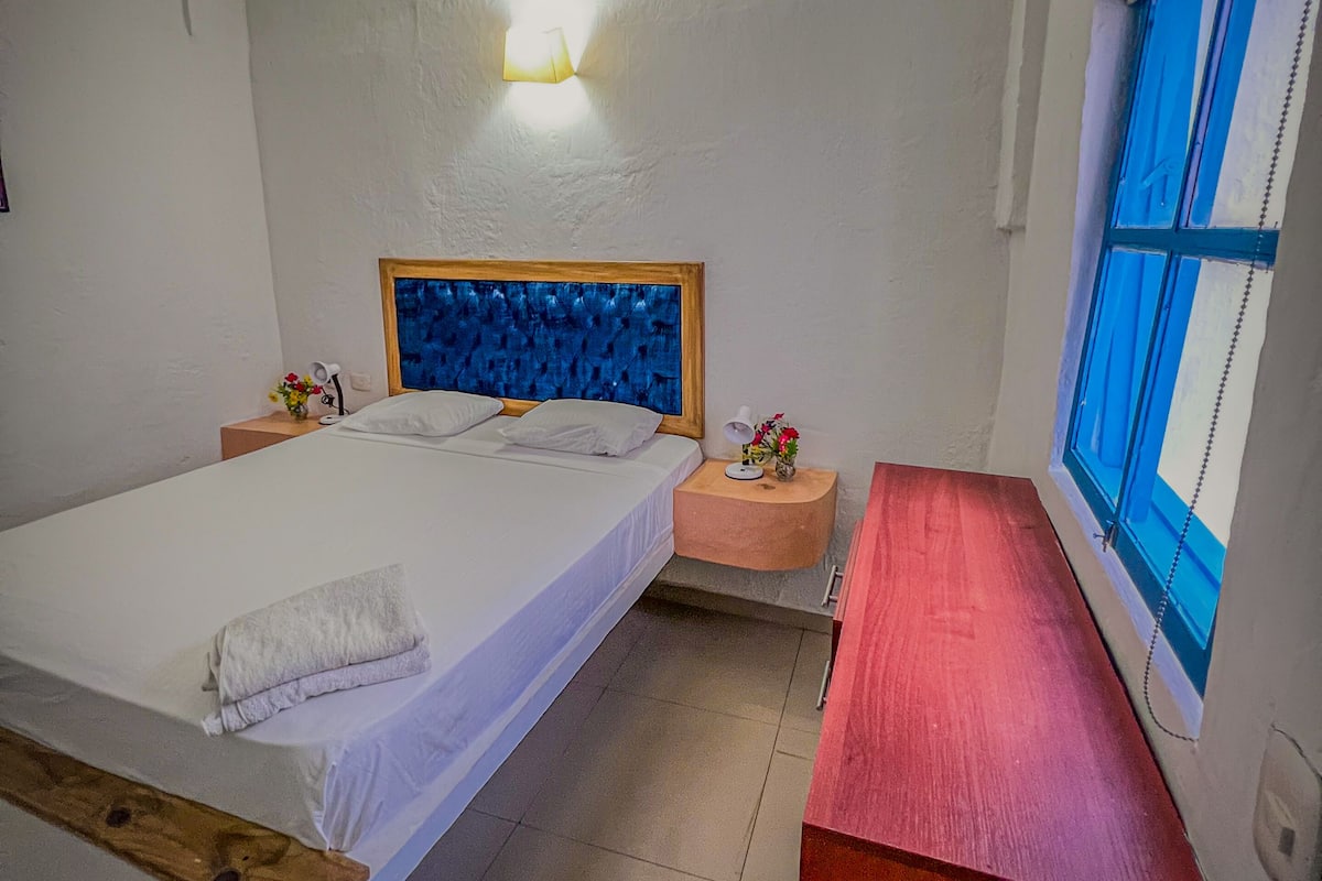 Ein Schlafzimmer mit einem großen Bett, Nachttischen, einer Ablage, einer roten Bank, einer Wandleuchte und einem Fenster mit blauen Vorhängen.