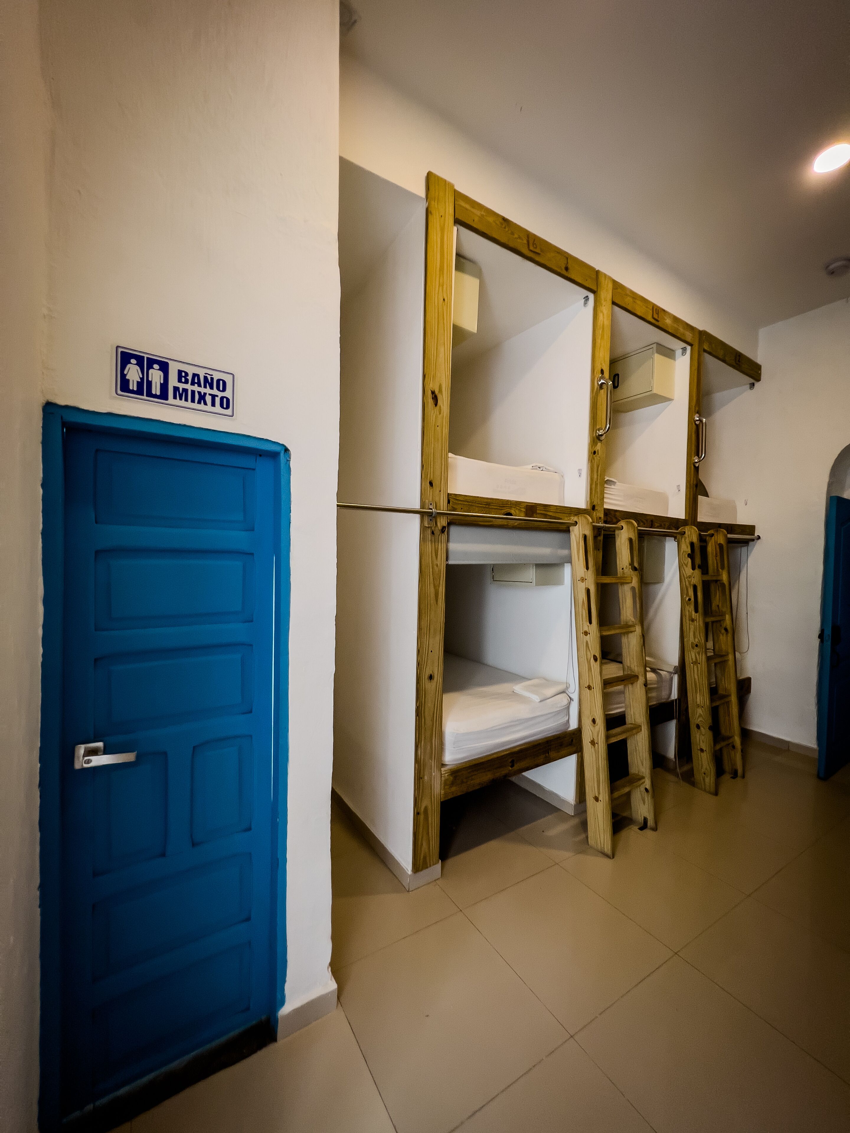 Una habitación con una puerta azul, una litera con escalera de madera y un cartel que indica baños para hombres y mujeres.