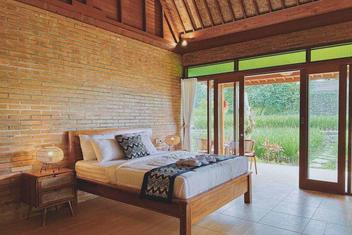 A bedroom with a brick wall, wooden ceiling, and a large window with a view of greenery.