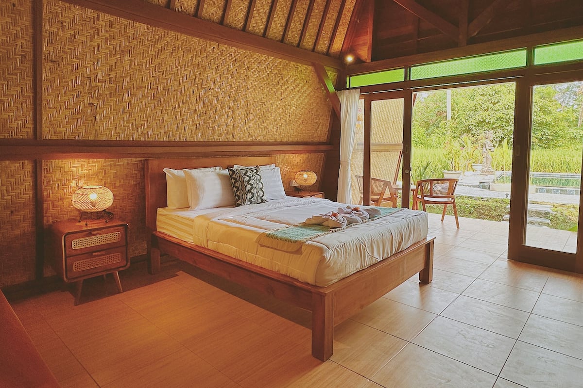 A spacious bedroom with a large bed, wooden headboard, and a woven wall. There is a small bedside table with a lamp and a chair outside the room.