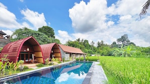 A pool with a tiled edge, surrounded by a lush green field. - Uma Raos Cottage (Ubud)