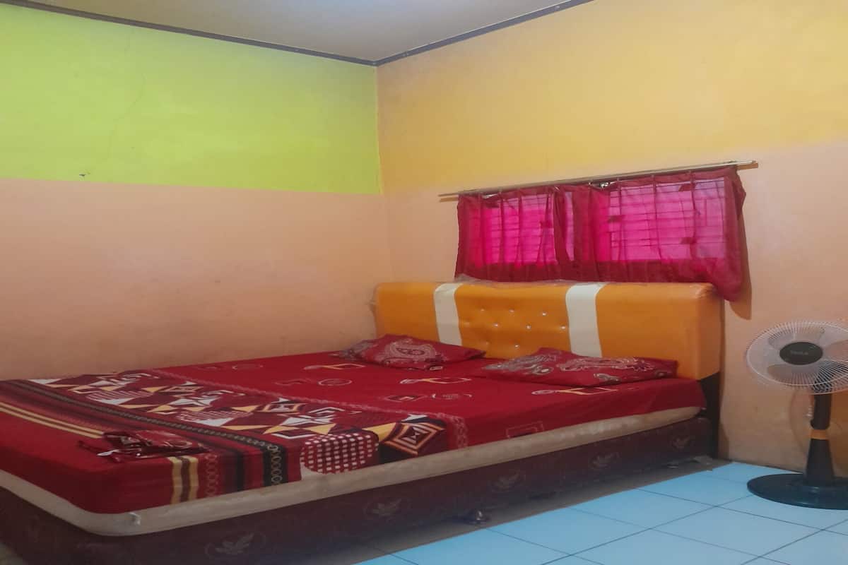 A small, single-bed room with a red bedspread, a window with red curtains, and a standing fan.