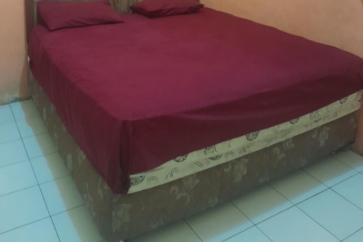 A single bed with a wooden headboard and a maroon bedspread in a room with peach walls and a window with blue curtains.