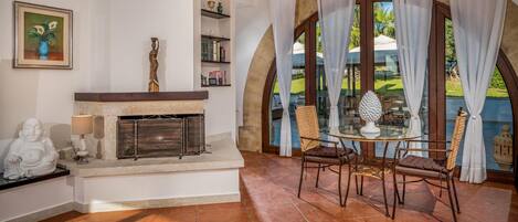 A cozy living room with a fireplace, a dining area with a glass table, and a view of a garden through large windows.