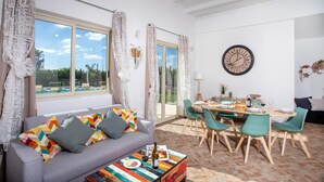 A modern living room with a grey sofa, colorful cushions, a dining table set for four, and a large wall clock. - Profumo di Zagara 6 2 Emma Villas (Ispica)