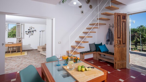 A modern living room with a glass dining table, a wooden bench with cushions, a staircase with wooden steps, and a view of the outdoors.