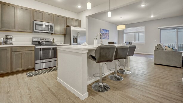 A modern kitchen with stainless steel appliances, a central island with bar stools, and a dining area with a gray sofa. - Top Flight (Colorado Springs)