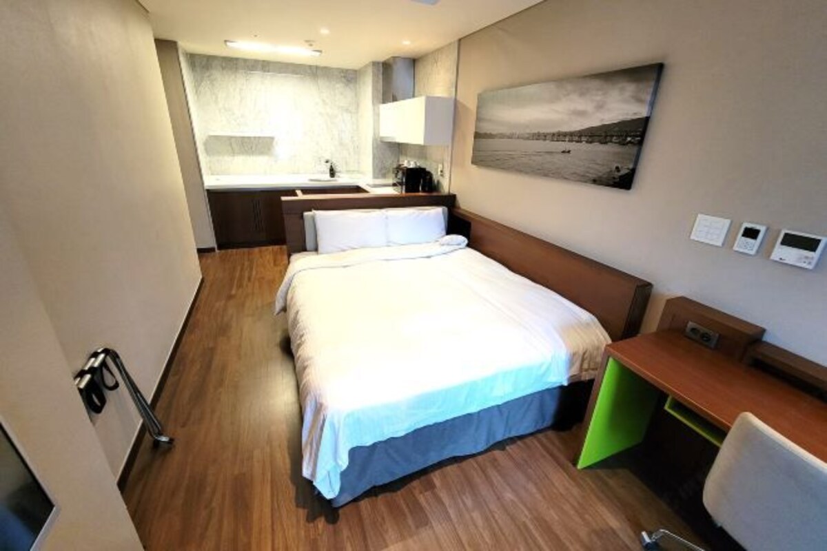 A hotel room with a bed, a desk, a chair, a small kitchenette, and a bathroom.