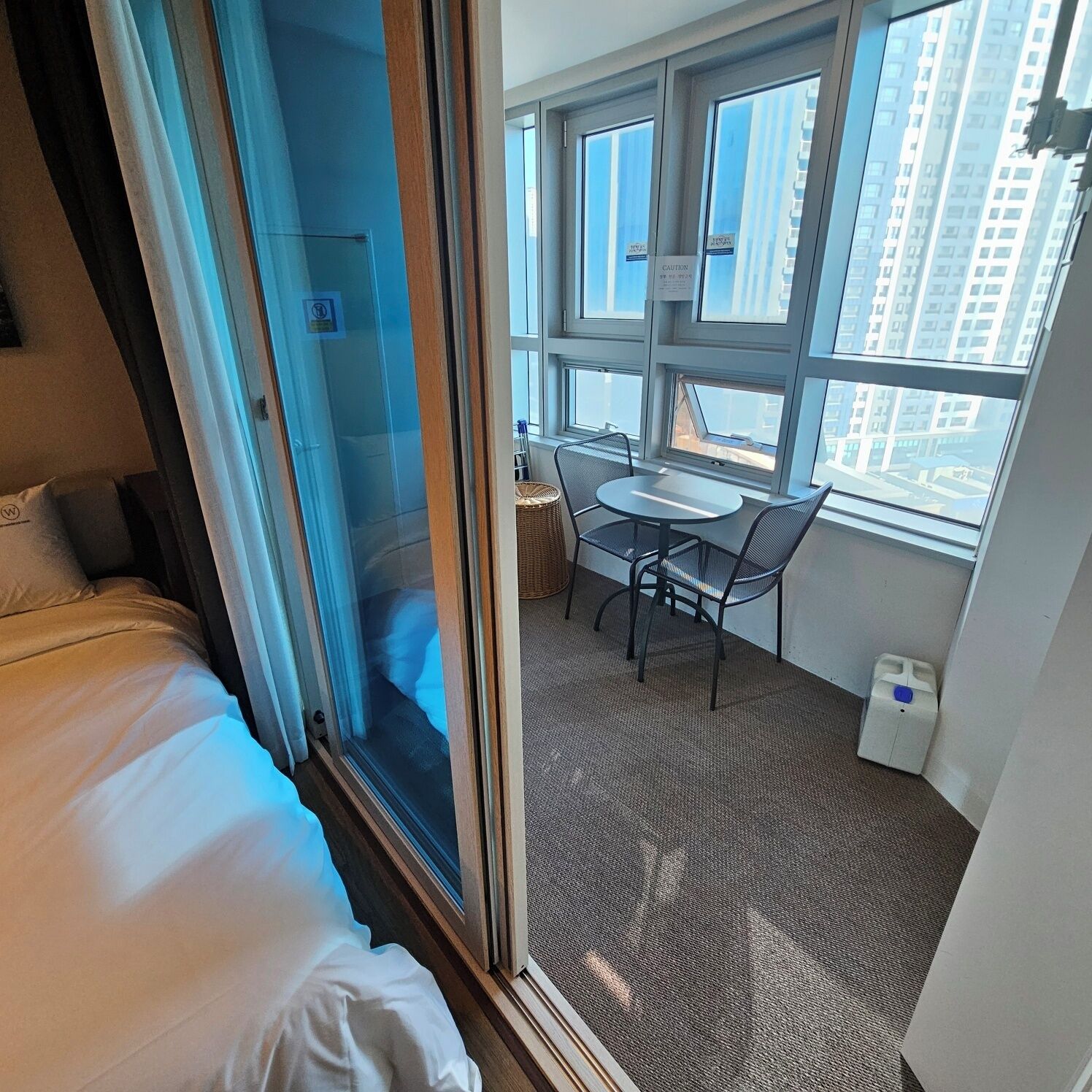 Habitación de hotel con cama, ventana con vistas a edificios altos, una mesita con sillas y un cesto de basura.