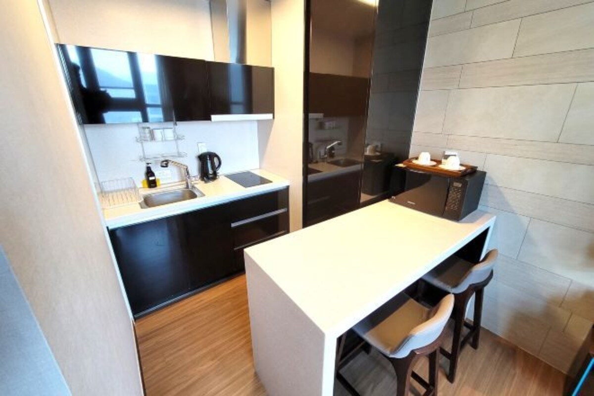 A compact kitchen with a white countertop, dark cabinetry, a coffee machine, and a small bar area with stools.