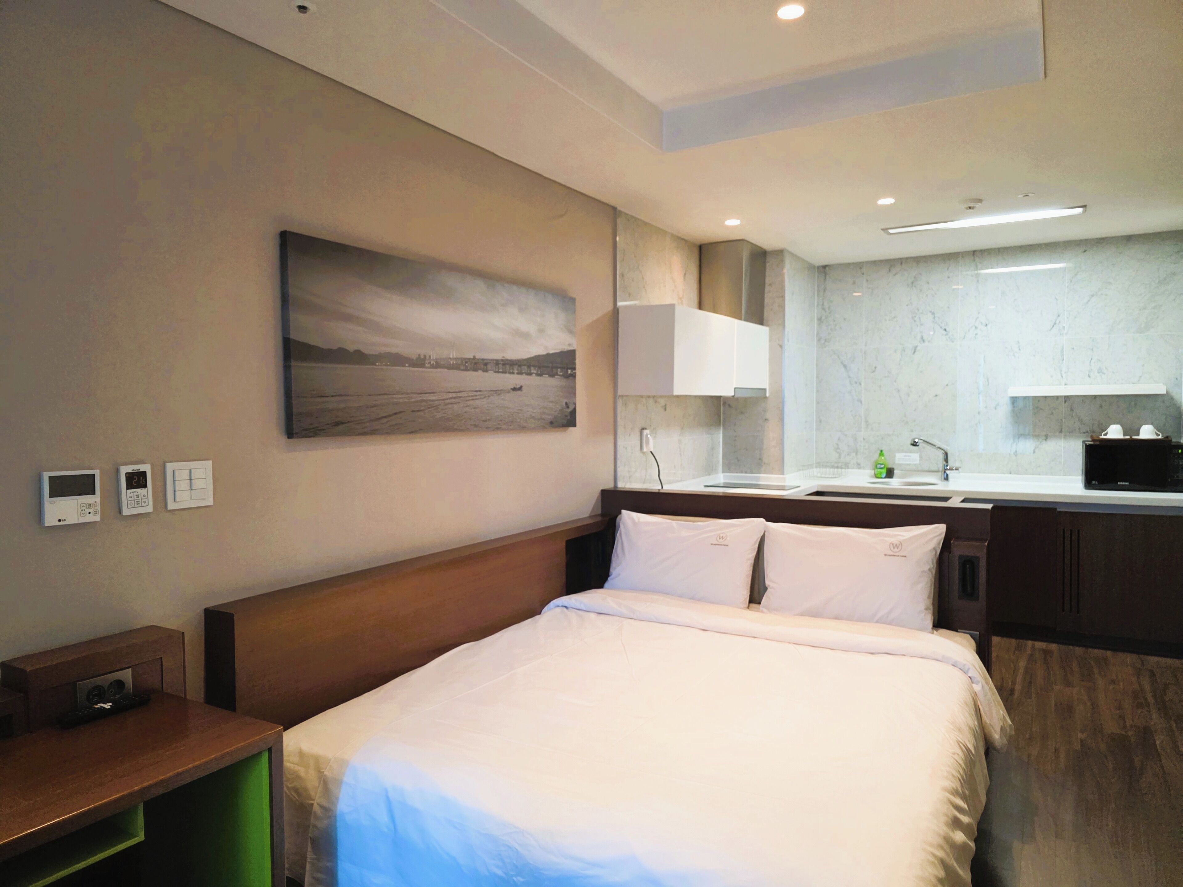 A hotel room with a bed, a bedside table, a nightstand, a wall-mounted thermostat, a wall-mounted clock, a painting of a landscape, a bathroom with a sink, a mirror, a countertop, a microwave, and a shelf.