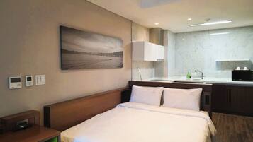 A hotel room with a bed, a bedside table, a nightstand, a wall-mounted thermostat, a wall-mounted clock, a painting of a landscape, a bathroom with a sink, a mirror, a countertop, a microwave, and a shelf.