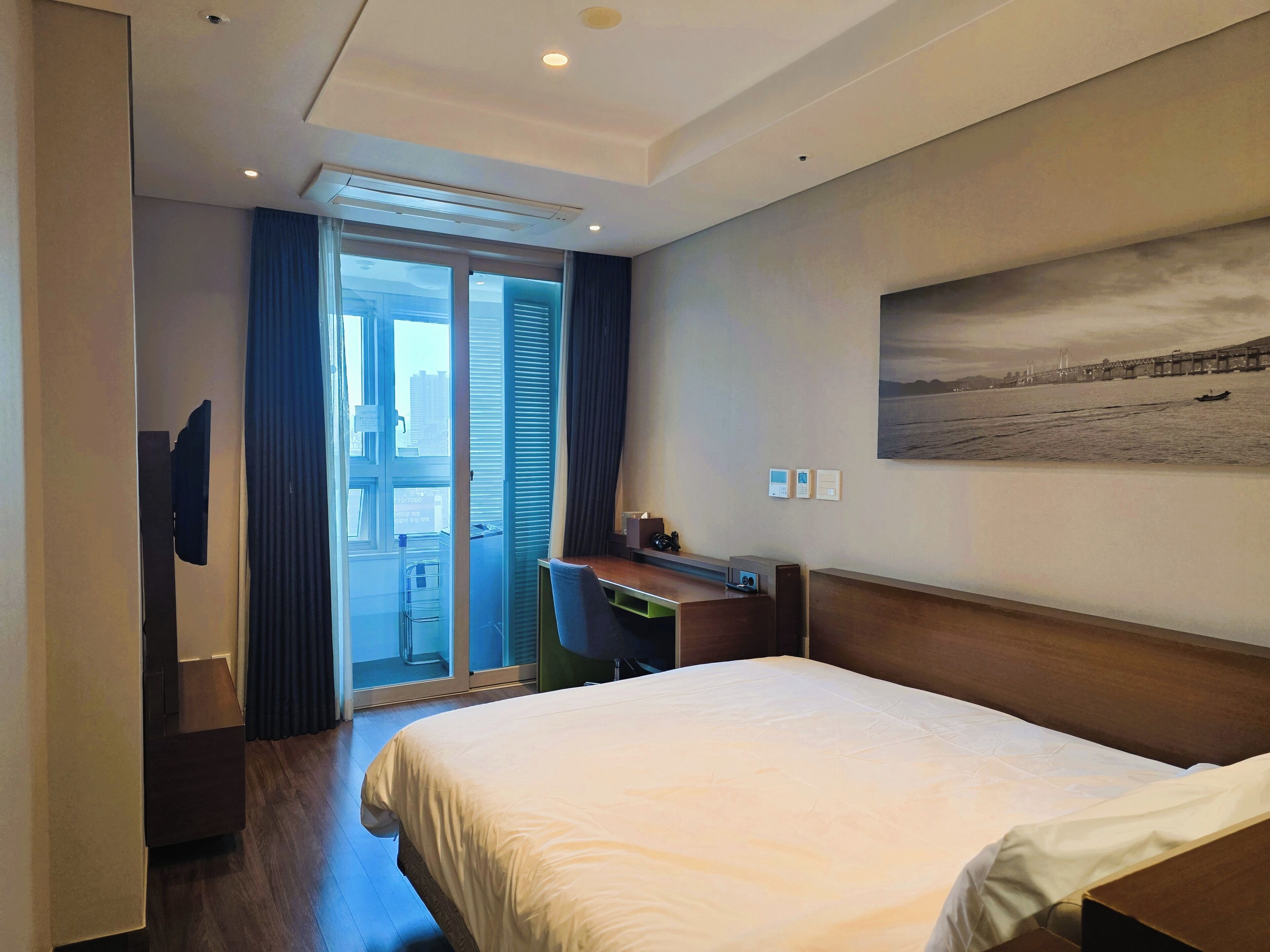 A hotel room with a bed, a desk, a TV, and a view of a cityscape through the window.