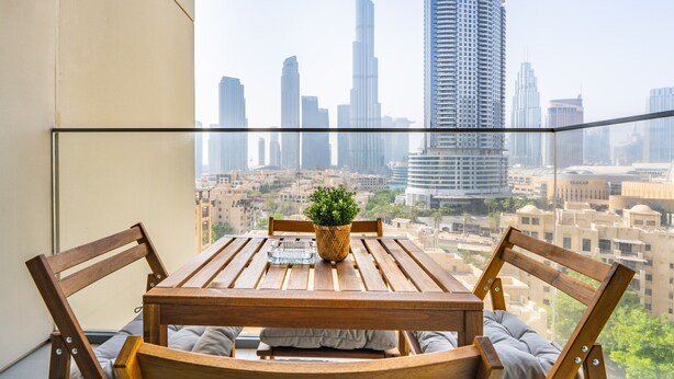 A balcony with a wooden table and chairs, a potted plant, and a view of the Dubai skyline including the Burj Khalifa.