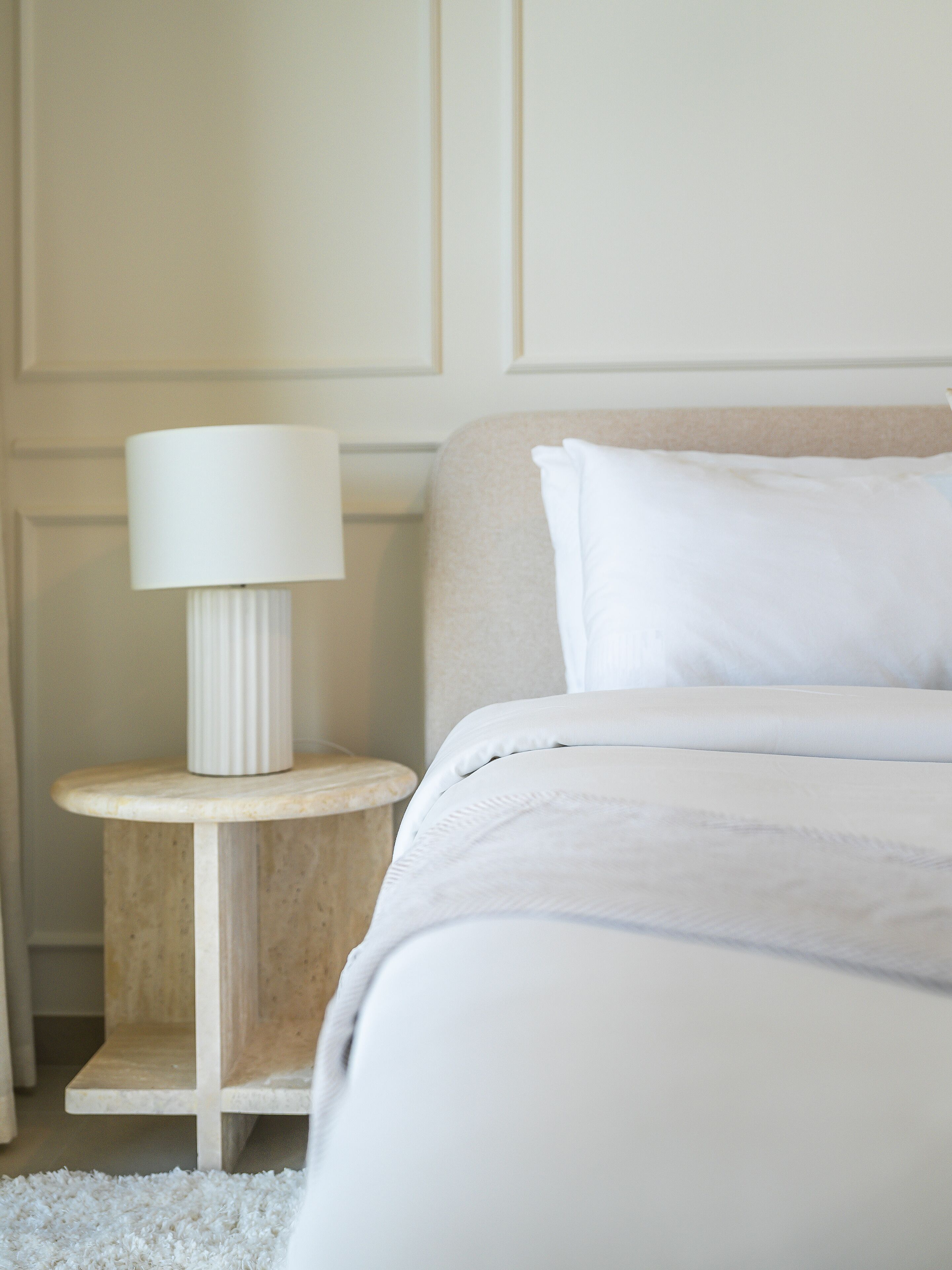 A neatly made bed with white bedding, a round wooden nightstand with a white lamp, and a beige wall with paneling.