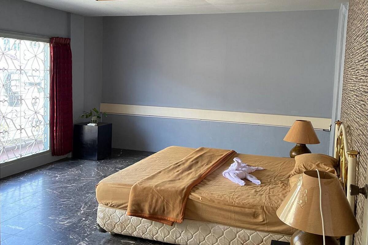 Un lit bien fait, avec une couverture beige et un oreiller, une lampe de chevet et une fenêtre ornée d’un rideau rouge.